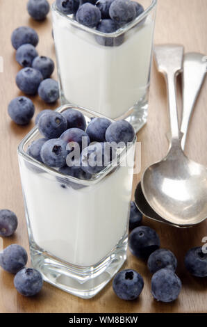 Yogurt biologici e i mirtilli in una piazza colpo di vetro e il cucchiaio sul tavolo di legno Foto Stock