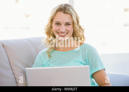 Ritratto di sorridente bionda seduta sul lettino usando computer portatile in salotto luminoso Foto Stock