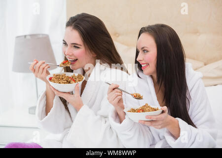 Abbastanza amici indossando accappatoi mangiare cereali a casa in camera da letto Foto Stock