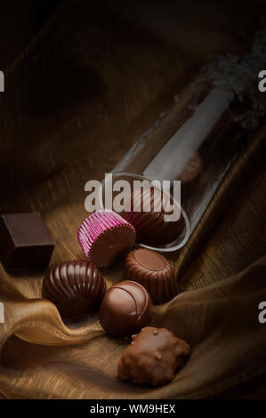 Fatti a mano di cioccolatini di lusso di diverse forme e sapori in un tubo di vetro riversandosi su tessuto morbido concettuale del regalo romantico con copyspace Foto Stock