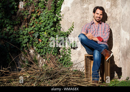 Sorridente modello maschile seduto sulla cassa di legno con le gambe incrociate Foto Stock