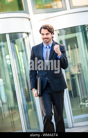 Motivati bel giovane imprenditore di lasciare il suo ufficio edificio con una valigetta sotto il suo braccio punzonatura il pugno in aria e sorridente dopo una suc Foto Stock