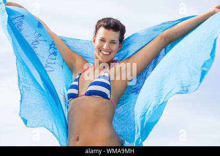 Carefree bella giovane donna in un bikini in esecuzione e saltando su una spiaggia con un bel blu turchese sciarpa galleggiante in aria dietro di lei come lei Foto Stock