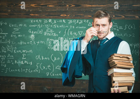 Bionda e giovane professore di mettere su i suoi occhiali. Uomo intelligente in tuta holding pila di libri Foto Stock