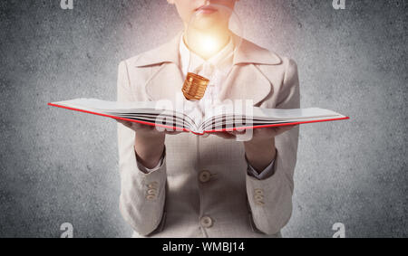 La donna che mostra la luce brillante lampadina sul libro aperto. Luce incandescente lampadina come simbolo idea creativa generazione. La donna in bianco business suit su sfondo di gr Foto Stock