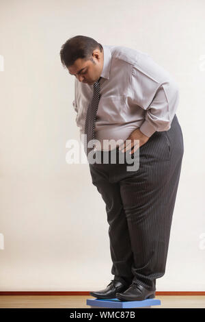Obeso di indossare abiti formali controllando il suo peso sulla macchina di pesatura Foto Stock