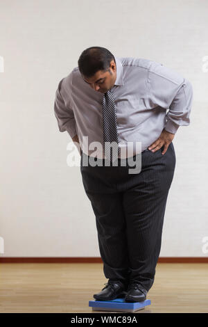 Obeso di indossare abiti formali controllando il suo peso sulla macchina di pesatura in piedi con le mani sulla vita Foto Stock