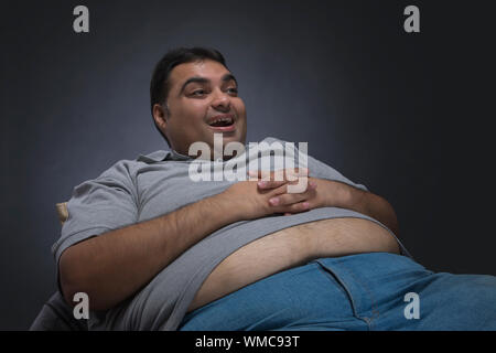 Uomo obesi con le mani sul suo ventre visibile al di fuori della sua maglietta seduto su una sedia che guarda lontano e sorridente Foto Stock