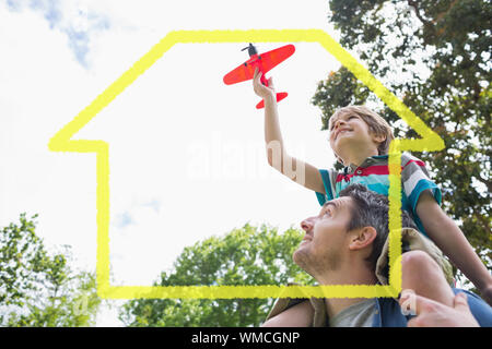 Ragazzo con aeroplano giocattolo seduto sulle spalle dei padri contro il contorno della casa Foto Stock