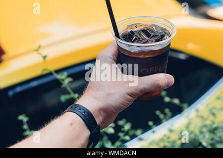 Gli uomini la mano che regge una tazza di plastica di ghiaccio nero caffè con una cannuccia e un taxi arancione di sfondo - non è di nessun aiuto di plastica il problema dell'inquinamento Foto Stock