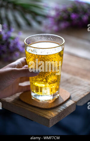 Tenere a mano un bicchiere di bevanda a base di semi di chia dorati su un tavolo di legno con fiori viola sullo sfondo. Foto Stock