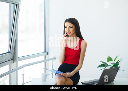 Affari. Ritratto di una giovane donna che tiene una mano di cartella blu. Vestita con T-shirt rossa e gonna nera. Donna di affari seduto sui tavoli nel Foto Stock
