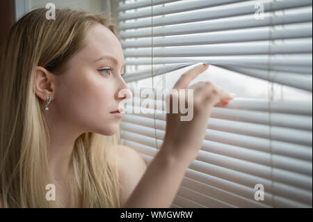 Giovane donna la separazione di lamelle di persiane e guardando attraverso la finestra. Foto Stock