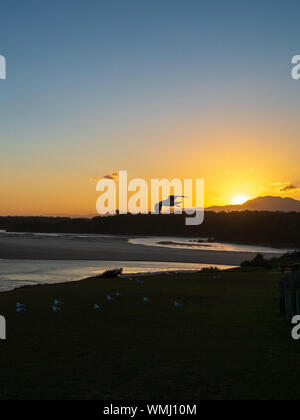 Un gabbiano silhouetted che vola contro un tramonto dorato mentre gli altri gabbiani d'Argento si radunano a terra in ombra al crepuscolo, Sawtell Australia Foto Stock