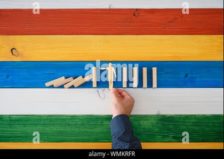 Vista superiore del maschio mettendo mano carta tagliata silhouette di un uomo di intervenire e impedire il domino di collassare. Su sfondo colorato della tavola di legno Foto Stock