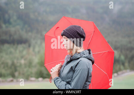 La donna in piedi che guarda pensieroso con un ombrello rosso sotto la pioggia Foto Stock