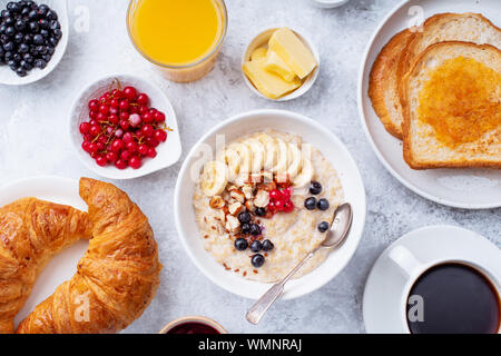 La prima colazione con i fiocchi d'avena con frutti di bosco e i dadi, croissant, fette biscottate con marmellata e burro, caffè e succo d'arancia Foto Stock