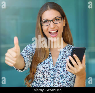 Gioioso di successo business donna con il pollice in alto contiene smart phone e guarda la fotocamera. Foto Stock