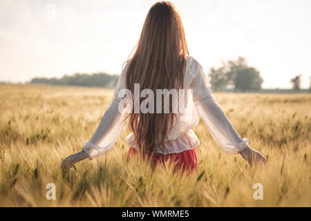 I Capelli rossi ragazza in un campo di grano al tramonto. Bella donna nel campo oro al tramonto, Retroilluminato toni caldi Foto Stock