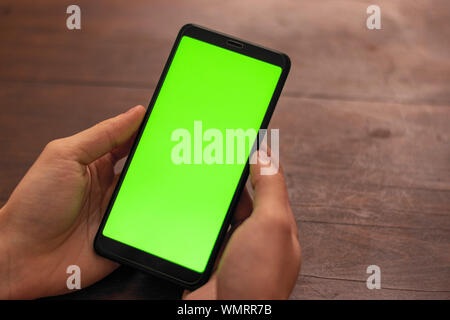 La mano della persona caucasica tenendo un nero smartphone non identificato con schermo verde su un tavolo di legno sfondo Foto Stock