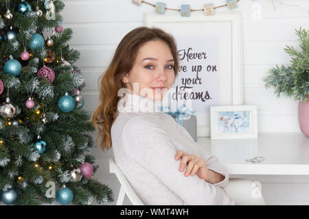 Ritratto di giovane sorridente donna seduta guardando la fotocamera su bianco muro di mattoni e albero di Natale sfondo Foto Stock