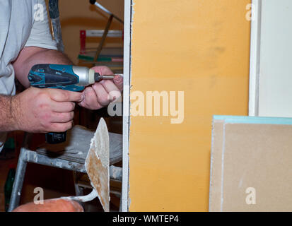 Maschio di mani tenendo la vite elettrico conducente durante il fissaggio di una parte di un pannello di cartongesso a parete durante lavori di ristrutturazione in un appartamento Foto Stock