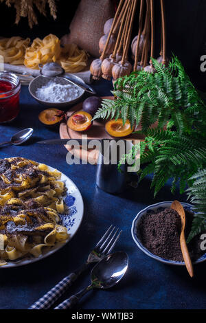 Pasta dolce dessert, tagliatelle con semi di papavero, prugna composta di prugne fresche, sfondo scuro, tavolo blu decorato con fiori freschi. Tutto secco popp Foto Stock