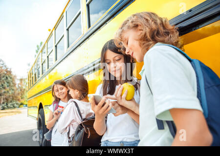 Un gruppo di compagni di scuola i bambini che vanno a scuola in autobus in piedi parlando di mangiare apple navigare in Internet sullo smartphone sorridente curioso close-up Foto Stock