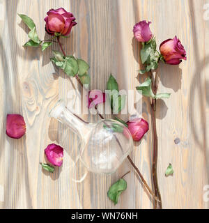 Tre secchi rose rosa, sparsi petali di fiori, foglie verdi vaso di vetro su sfondo di legno top view close up, vintage romantiche decorazioni floreali Foto Stock