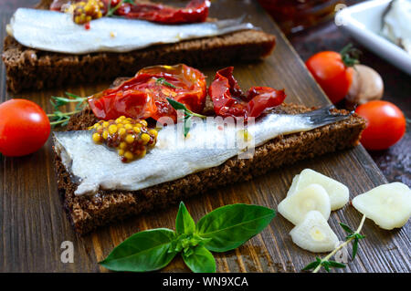 Gustosa smorrebrod su una tavola di legno. Panini con il nero del pane di segale, pomodori secchi, acciughe salate, la senape. Foto Stock