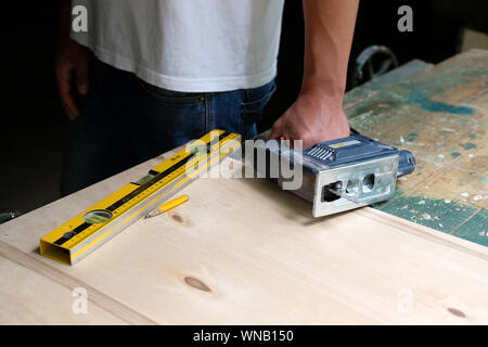 Le mani di umana holding seghetto alternativo elettrico giacente su un banco di lavoro con bolla di livello e penna gialla. Concetto di falegnameria. Foto Stock