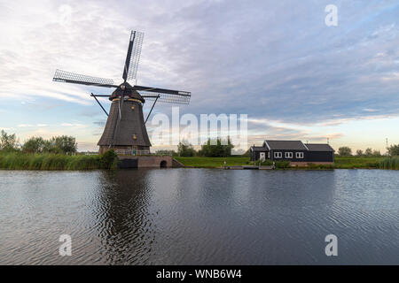 Mulino a vento olandese recante lungo il canale con erba selvatica soffiata dal vento forte all'inizio del momento al tramonto Foto Stock