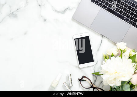 La donna lo spazio di lavoro con computer portatile, peonia bouquet di fiori, smartphone, accessori, occhiali su marmo bianco dello sfondo. Lay piatto home office desk. T Foto Stock