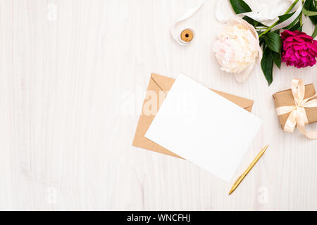 Piatto peonie laici fiori, artigianato, busta vuota scheda carta, confezione regalo, penna, nastro su sfondo di legno. Vista superiore dello spazio di lavoro femminile, wedding invitatio Foto Stock