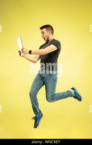 A piena lunghezza Ritratto di felice saltando l uomo con i gadget isolato su sfondo giallo. Le moderne tecnologie, libertà di scelte concetto, emozioni concetto. Utilizzando laptop e smartwatches in volo. Foto Stock