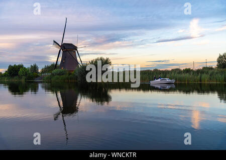 Mulino a vento olandese recante lungo il canale con erba selvatica soffiata dal vento forte all'inizio del momento al tramonto Foto Stock