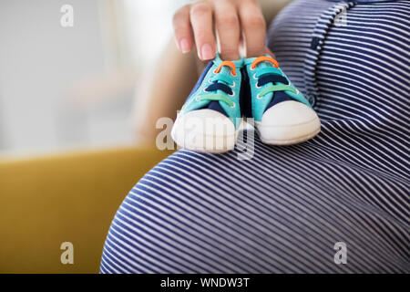 Close up della donna incinta seduti sul divano di casa azienda scarpe per bambini su stomaco Foto Stock