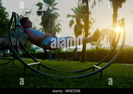 Uomo caucasico in appoggio in una amaca tra alberi di palma e guardare il tramonto. Foto Stock