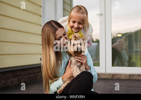 Bella donna con capelli lunghi biondi holding cane piccolo Yorkshire terrier outdoor, prossima seduta bambina. Felice madre e figlia con pet sul Foto Stock