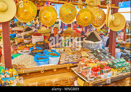 KAKKU, MYANMAR - 20 febbraio 2018: il piccolo box del mercato agricolo con molti prodotti locali - gli agricoltori hets conica, cumuli di aglio, pacchetti di Foto Stock