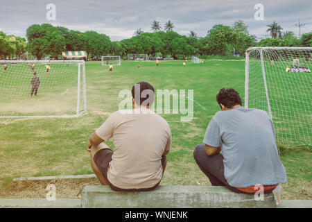 Vista posteriore dei genitori maschili acclamava i loro bambini a giocare a calcio nella scuola. Foto Stock