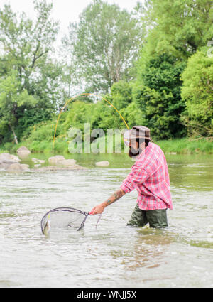 Barbuto brutale fisher la cattura di pesci trote con net. Se regolarmente del pesce si sa come gratificante e rilassante è la pesca. La pesca è un sorprendente accessibile ricreativo sport all'aperto. La pesca hobby. Foto Stock