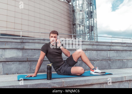 Atleta maschio, estate in città, in appoggio su una camicia di yoga, chiamando al telefono, di indossare abbigliamento sportivo pantaloncini e una T-shirt, un agitatore con acqua e proteina, un Foto Stock