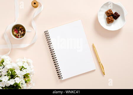 Romantico home office desk con blocco di carta, daisy fiori, tazza di tè, candy, nastro pastello su sfondo rosa. Appartamento laico, vista dall'alto, overhead. Foto Stock