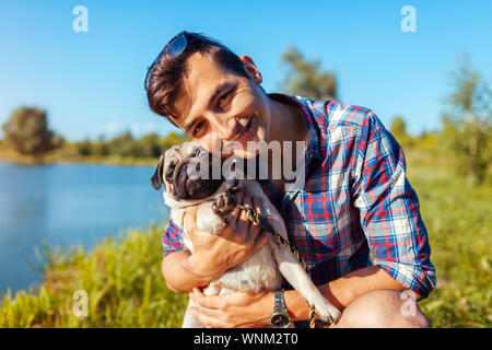 L'uomo abbracciando pug dog nel parco. Felice l'uomo a piedi con PET da parte di estate lago Foto Stock