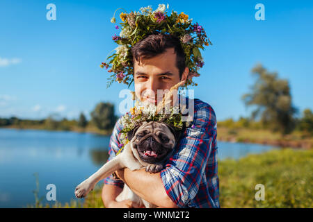 Uomo con pug dog con la ghirlanda di fiori sulla testa. Felice l'uomo a piedi con PET da parte di estate lago Foto Stock