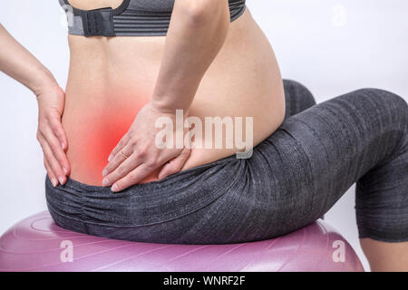 Dolori e. Attiva donna incinta esercitarsi nel centro fitness in sala yoga. La giovane madre prevedente tenendo le mani sul retro e sitts al fitness Foto Stock