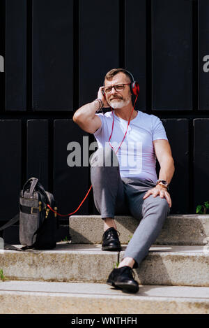 Barbuto uomo maturo seduto sui gradini con le cuffie durante i momenti di relax in una giornata di sole Foto Stock
