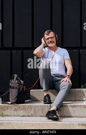 Barbuto uomo maturo seduto sui gradini con le cuffie durante i momenti di relax in una giornata di sole Foto Stock