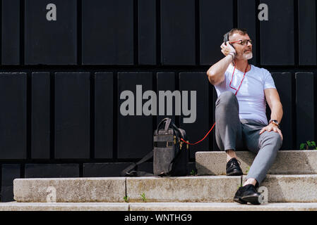 Barbuto uomo maturo seduto sui gradini con le cuffie durante i momenti di relax in una giornata di sole Foto Stock